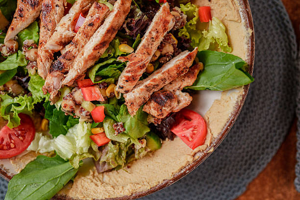 A bowl of colorful grilled chicken salad featuring mixed greens, cherry tomatoes, corn, and a light dressing. The dish is served on a wooden table in a cozy café setting, showcasing a healthy and vibrant meal perfect for lunch or a balanced diet theme.
