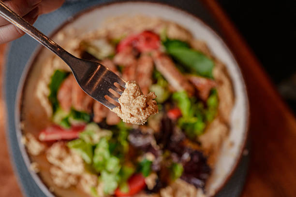 A close-up of a fork lifting a bite of creamy hummus mixed with fresh greens and grilled chicken from a Mediterranean-style bowl. The blurred background emphasizes the texture and detail of the food, evoking a sense of freshness and appetizing simplicity.