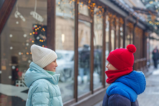 Boy and girl wearing warm jackets and hats standing face to face and chatting outside a festive Christmas-themed storefront in winter.