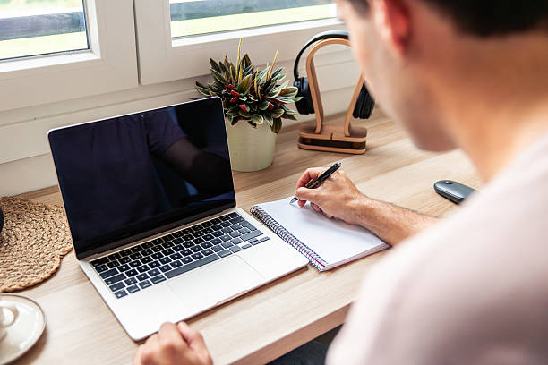 istock Young man teleworking remotely and writing notes at his home office desk 2240809831