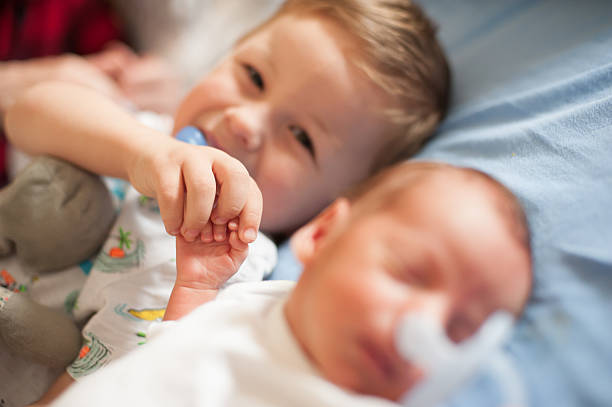 In a cozy bedroom, an older baby brother gently holds his newborn sibling's hand as they both sleep soundly. The warm atmosphere highlights their bond in this tender moment.