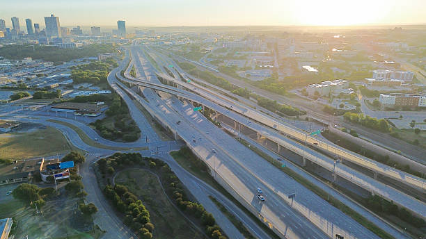 istock Curved stacked interchange channels traffic into Fort Worth downtown skyline, foreground has logistics hub, flat-roof warehouses, and directional ramps framed by early light and tree buffers 2240836310