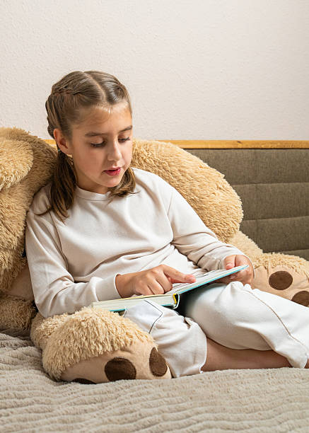 istock Young child sits on bed with a large stuffed bear, enjoying a book in a warm home setting. Ideal for content about childhood reading, relaxation, family atmosphere. 2240840027