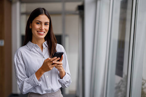 istock Professional businesswoman smiling while sending message on mobile phone, standing in modern office corridor 2240857249