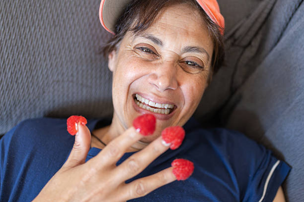 Mature woman laughing, balancing ripe red raspberries on her fingertips, enjoying a playful, healthy snack at home.carefree, content, and naturally joyful in a casual moment