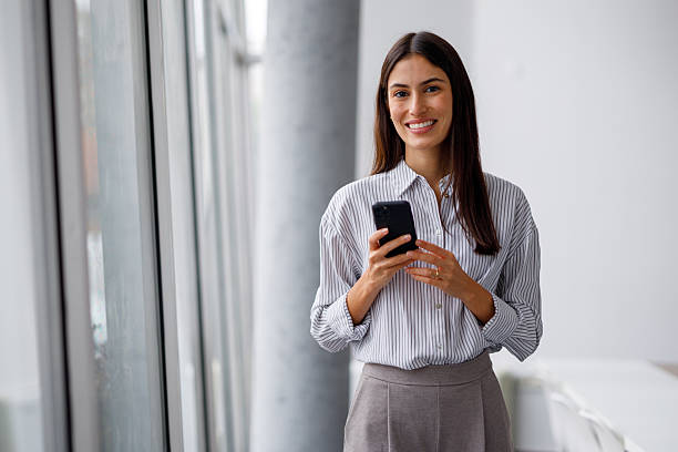 istock Woman standing in a bright office space, smiling and holding a mobile phone, representing professional connectivity 2240858839
