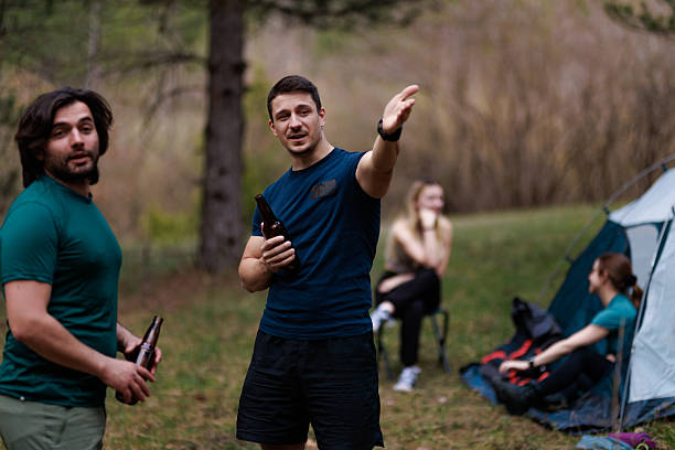 Groups of friends gather around a campsite in a beautiful forest, relaxing, chatting, and enjoying drinks as they bond over shared adventures and good times outdoors.