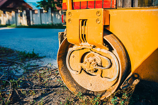 Yellow road roller with a large, rusty metal drum resting on a patch of grass and dirt. Freshly paved asphalt road is visible in the background, hinting at recent construction