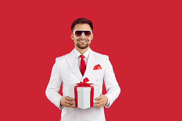 Joyful attractive young man in formal suit with gift box in hands isolated on red background. Caucasian man in white suit with red tie and glasses holds white gift box with red ribbon. Banner.