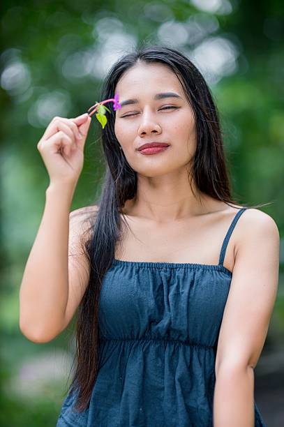 A young woman enjoys a tranquil moment in the forest, delicately holding a flower. The serene environment highlights her beauty and connection to nature.
