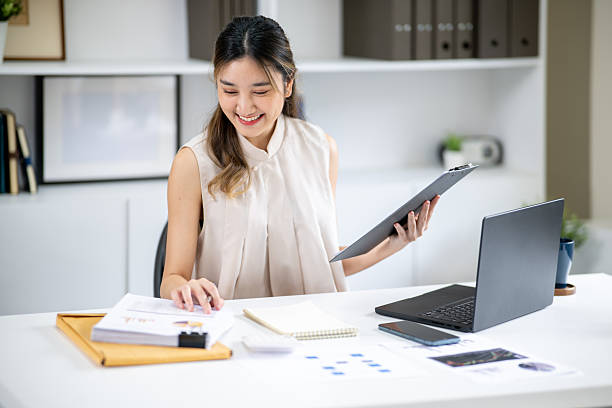 istock Smiling woman office worker holding clipboard as looking at document aside laptop at table in office 2240896114