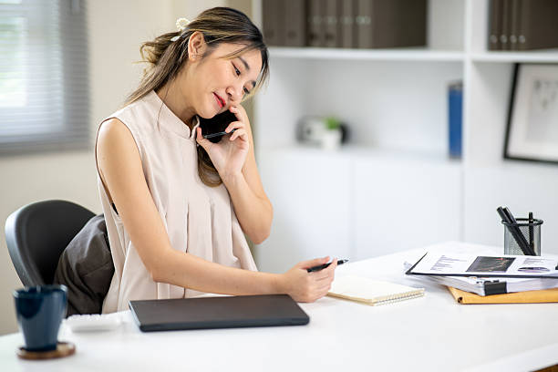 istock Pretty woman office worker holding pen looking at notebook while talking on phone at table in office 2240896649