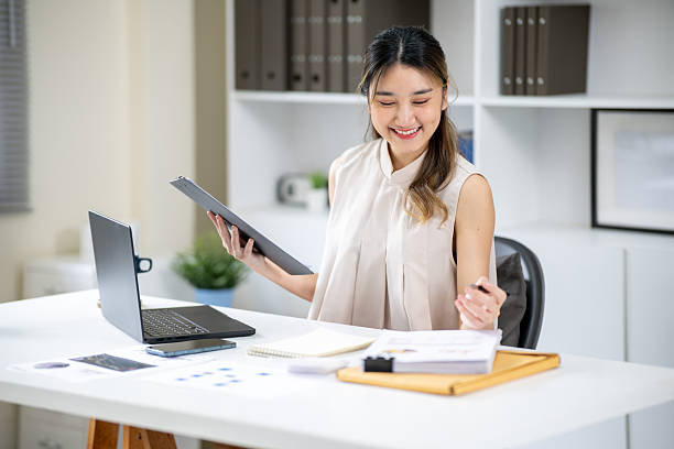 istock Woman office worker holding pen and clipboard as looking at document aside laptop at table in office 2240896678