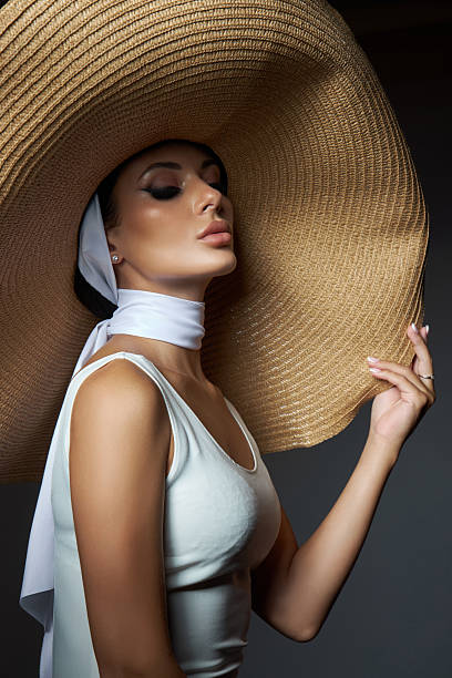 Model with refined makeup wears a large straw hat and a chic scarf, exuding elegance and sophistication in a dimly lit studio environment. photo captures her grace and poise