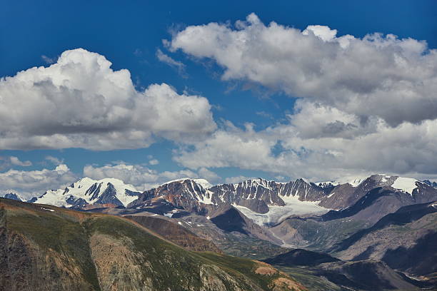 Snow-covered mountains rise majestically against a deep blue sky filled with fluffy clouds during the summer season. scene captures the beauty of nature in full splendor. Russia, Altai Mountains