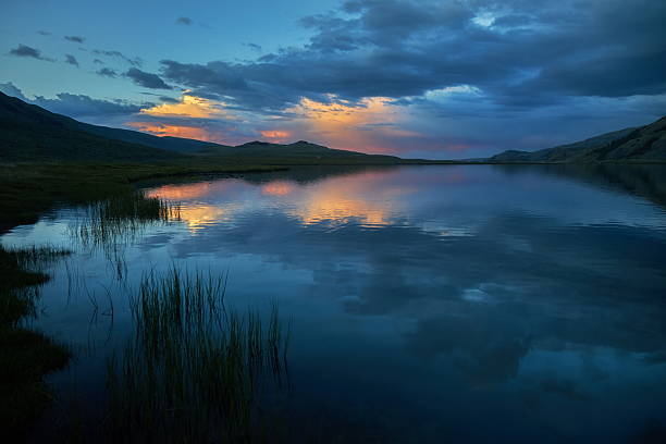 tranquil lake reflects colorful clouds at dusk, surrounded by distant mountains. calm water mirrors the stunning hues in the sky, creating a peaceful evening scene