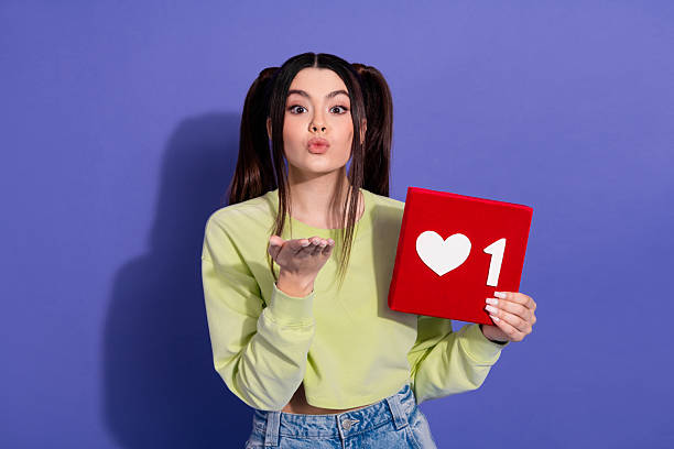 istock Young woman with casual style holding a heart icon while making a blowing kiss gesture against a purple background 2240920006