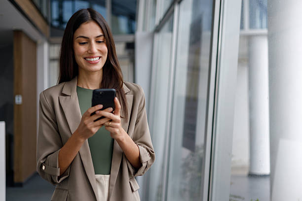 istock Businesswoman actively using a mobile phone, smiling while standing in a modern office building, connecting with technology 2240922557