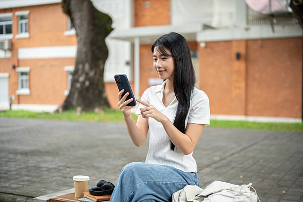 istock Young asian woman student holding looking at phone typing or making a call sits on bench by sidewalk 2240922719