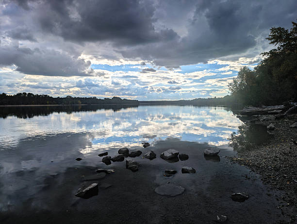 A wide-angle landscape captures a dark and expressive scene of the Danube River, where large, dramatic gray clouds and patches of bright blue sky are mirrored by the still, reflective water, while scattered dark rocks sit in the shallow, gravelly foreground.
