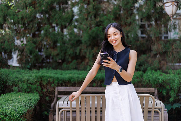 Young asian woman enjoys using her smartphone in a serene park setting, showcasing connectivity and modern lifestyle