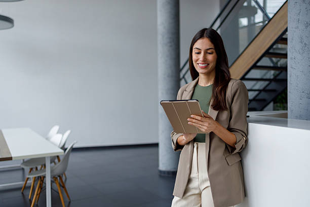 istock Businesswoman standing in modern office using digital tablet, smiling and managing work with finance technology 2240925578