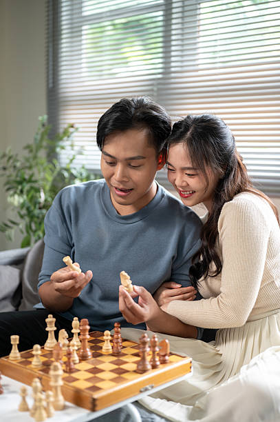 Asian man holding chess piece talking explaining to a woman hugging his arm aside sofa table at home. Husband and Wife, Board game, Bonding Activity.