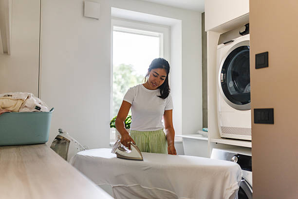 A young woman stands in a bright laundry room ironing clothes on an ironing board. A washing machine and laundry basket are visible, adding a sense of domestic life and responsibility.