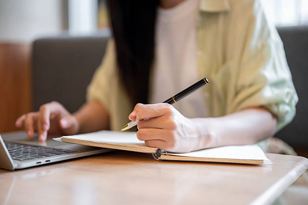 istock Close up woman student holding pen writing in notebook looking or typing on laptop at table in cafe. 2240927618