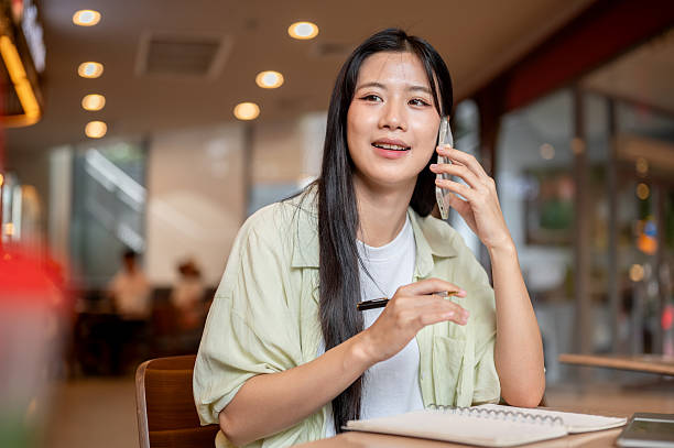 istock Pretty asian woman student holding pen over notebook while talking on phone at wooden table in cafe. 2240927742