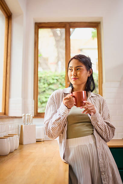 istock Contemplative Asian Woman with Coffee Mug Leaning on Kitchen Counter 2240928358