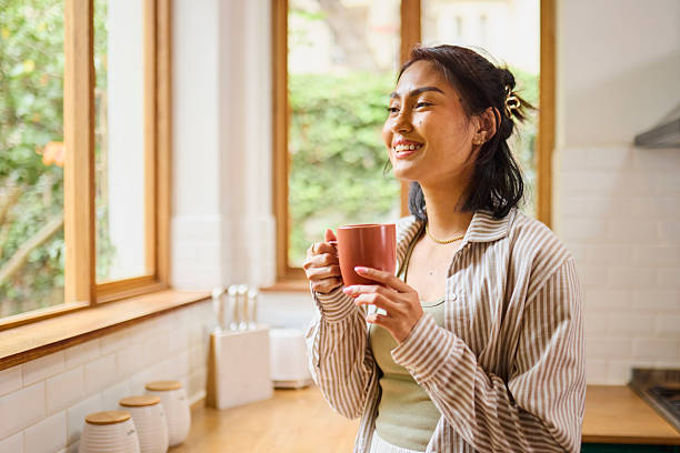 istock Happy Asian Woman Enjoying Coffee or Tea by Kitchen Window 2240928393