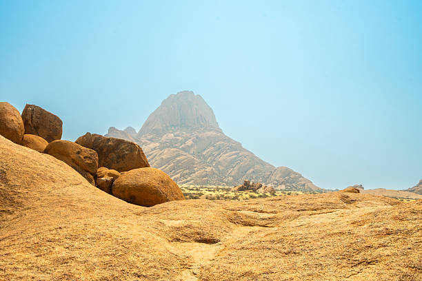 Granite peaks and rugged desert landscape at Spitzkoppe Community Campsite, Namibia. Golden grassland with scattered acacia trees contrasts against dramatic rocky mountains under a clear sky.