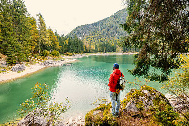 Man in red jacket with backpack standing on rocky shore overlooking clear green mountain lake. Outdoor adventure and nature exploration. Peaceful wilderness landscape with forest and water.