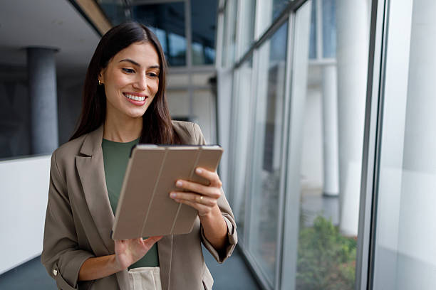 istock Businesswoman standing in modern office corridor using digital tablet, smiling and connecting with technology for corporate work 2240930973