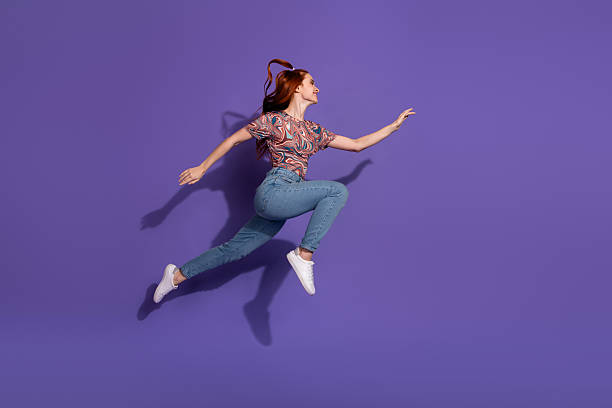 istock Energetic young red-haired woman leaping joyfully against a solid purple background, showcasing vibrant casual fashion and style 2240931765