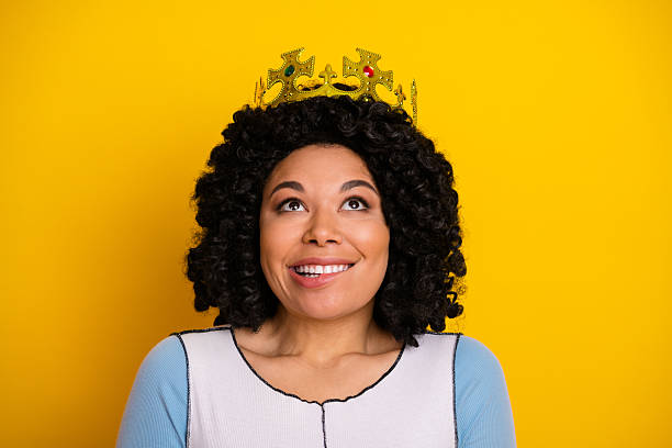 Portrait of a happy young woman wearing a golden crown, posing against a bright yellow background, exuding confidence.