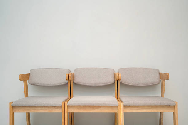 Three simple wooden chairs with light fabric cushions placed side by side against a clean white wall. The minimalist interior design represents calm, and simplicity in home or office waiting area.