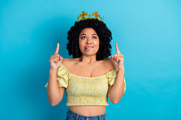 istock Young cheerful woman wearing a crown makes hand gestures while posing in a stylish outfit against a blue background 2240933045