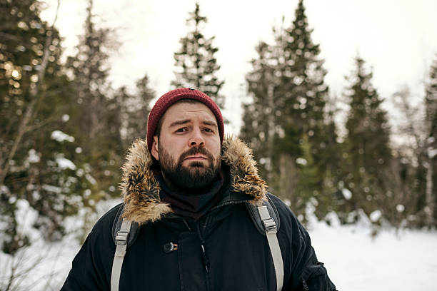 Portrait of adult bearded man in warm jacket and hat standing with backpack in winter woods looking at camera