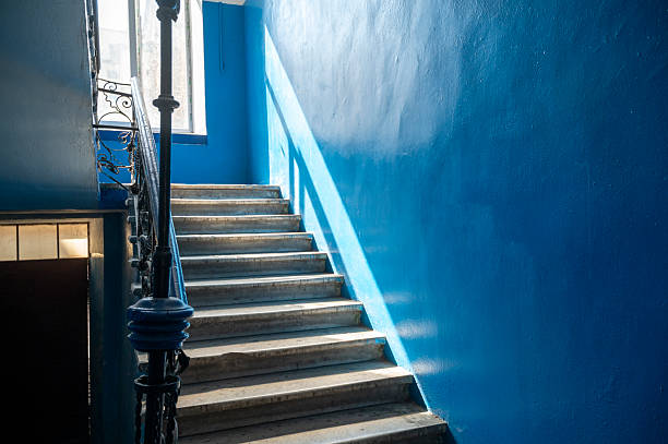 Marble staircase with openwork black iron railings and a bright blue wall, illuminated by the sun. Diagonal perspective of the staircase with interior lighting and a frame.