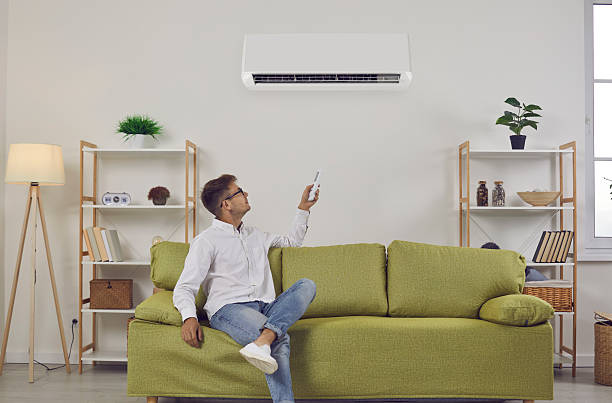 istock Man who's sitting on sofa at home turns on wall air conditioner using remote control 2240935905