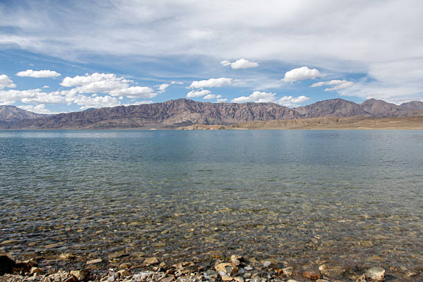 A lake with clear water surrounded by mountain range under a sky with scattered clouds, showcasing natural landscape.
