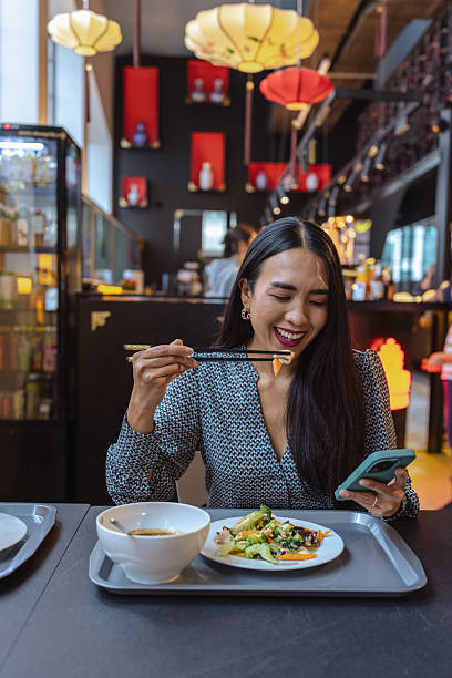Asian woman enjoying lunch at a modern restaurant while browsing her phone and tasting food