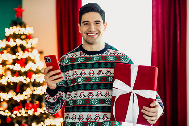 istock Young man celebrating Christmas holding a smartphone and present in a cozy holiday setting with festive decorations 2240942514