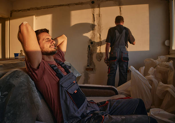 Two male construction workers in overalls inside a partially renovated room. One worker relaxes on a couch in warm sunlight while the other works on the wall. The scene shows a calm moment during renovation progress.