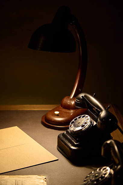 A carbolite lamp and a rotary telephone sit on the dark green desk. A folder with classified documents lies nearby