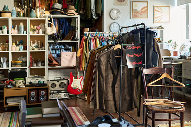 Vintage clothing hanging on racks in thrift shop surrounded by shelves filled with assorted accessories, musical instruments, and retro electronics, no people visible