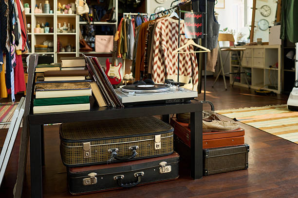Vintage suitcases, books, and record player displayed on wooden table in thrift shop with racks of assorted clothing and accessories visible in background, suggesting secondhand retail setting