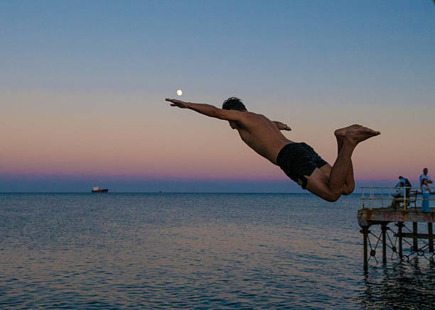 A young man performing a jump from a coastal pier into the Mediterranean Sea in Limassol, Cyprus, during the sunset and the moonrise.